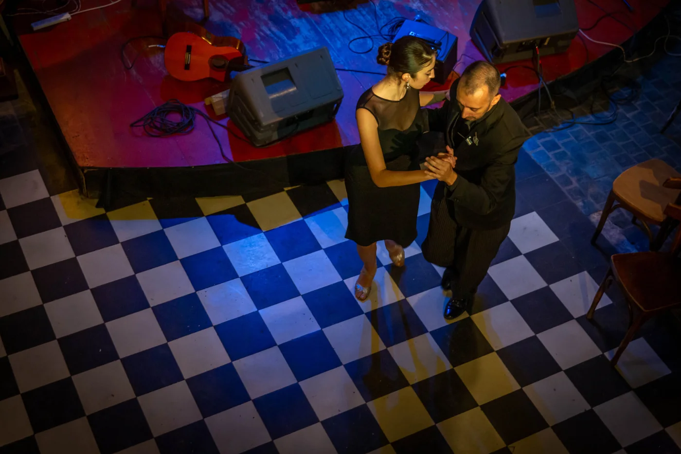 A tango couple dancing on a black-and-white checkered floor beside a small stage with a guitar and sound equipment (representative photograph of a Rioplatense tango show, not Primuseum's own room)